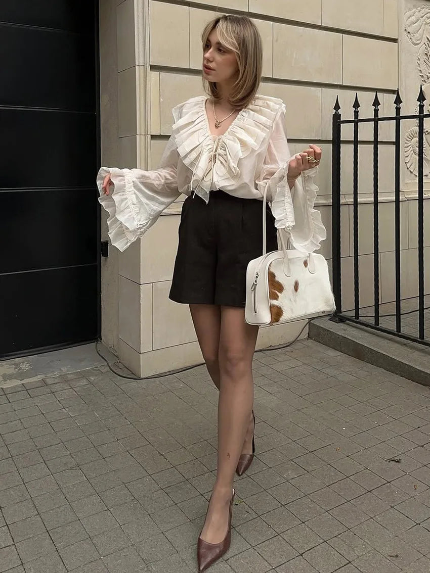 Woman in a white blouse with ruffles and black shorts holding a white and brown handbag on a city street.