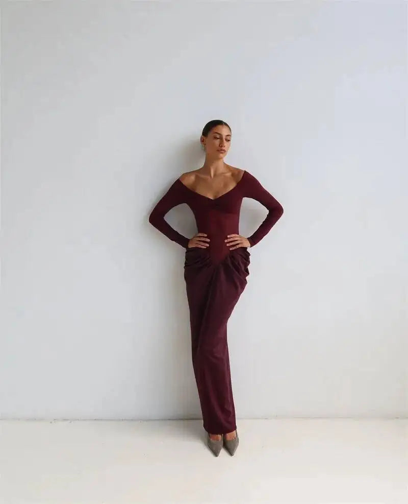 A person posing in an off shoulder long sleeve maxi dress with ruched waist detail against a white wall.