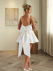 Woman in a white dress with a large bow standing in a room with wooden flooring and a neutral wall.
