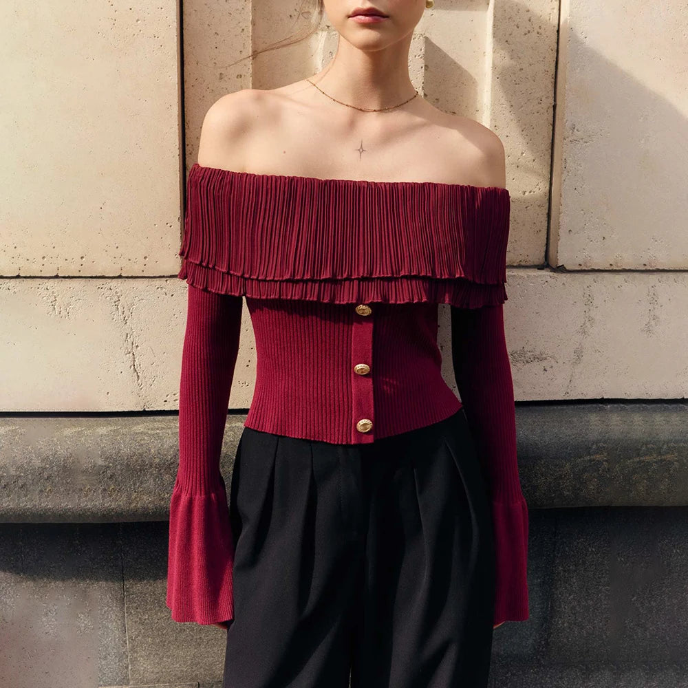 Person wearing a burgundy off-shoulder top with ruffled details against a stone wall.