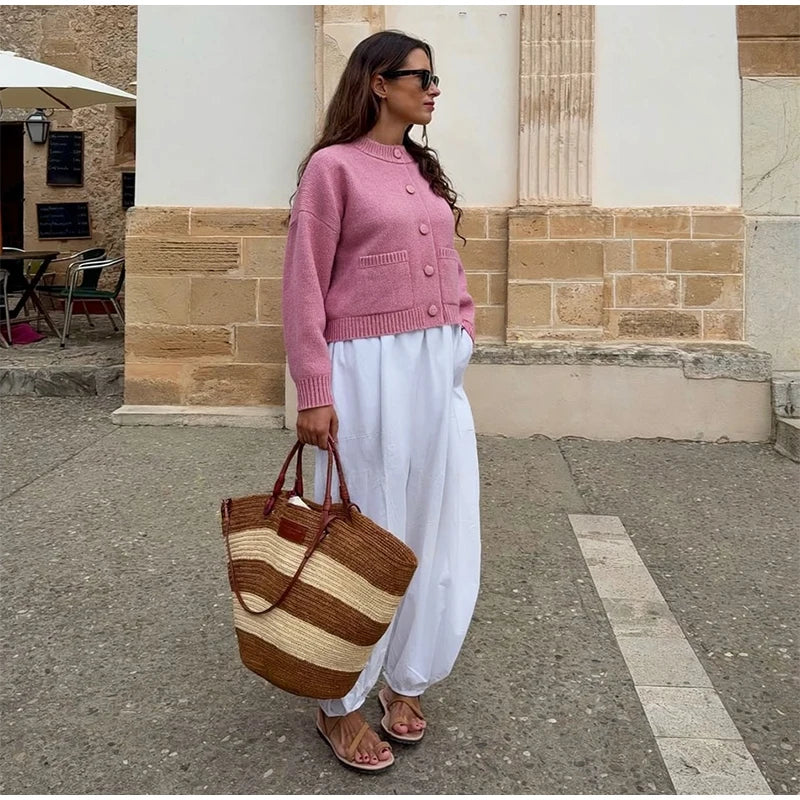 Woman in pink cardigan and white pants holding a striped bag on a street.