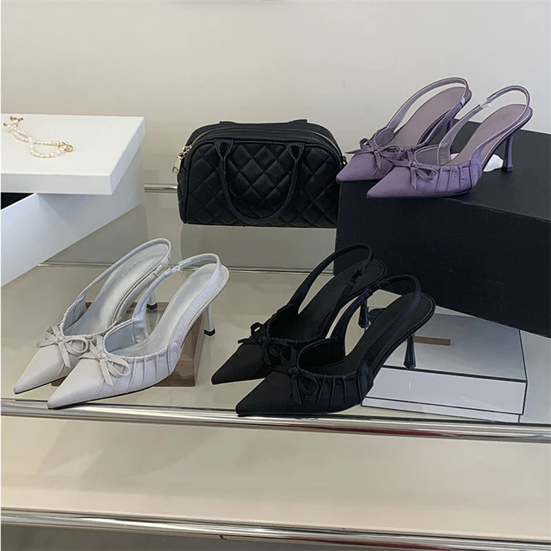Pair of white heeled shoes, black quilted handbag, and purple high-heeled shoes on a reflective surface.