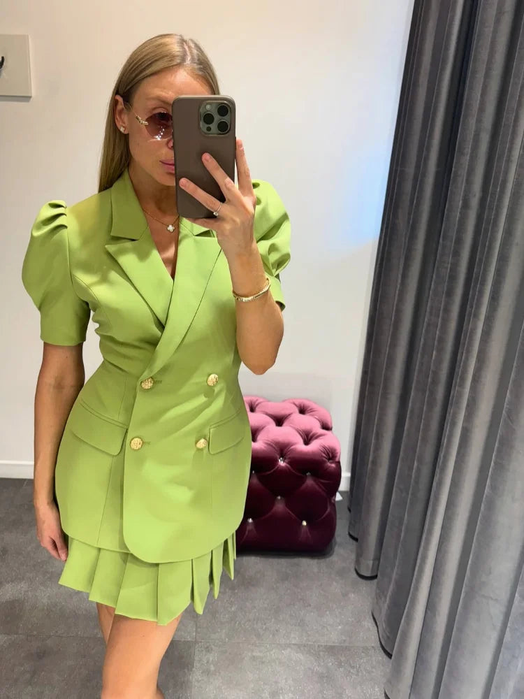 Person wearing a green outfit taking a mirror selfie in a room with a purple ottoman and gray curtain.