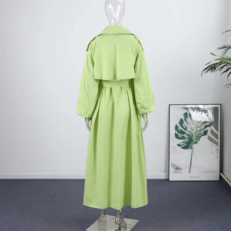 Mannequin wearing a light green trench coat in a room with a framed plant picture on the wall.