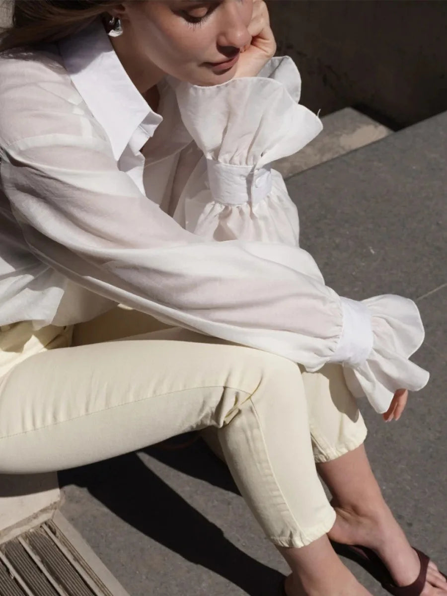 Person wearing a white blouse with ruffled sleeves and light-colored pants sitting on a bench.