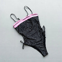Flat lay of a black and white polka dot one-piece swimsuit with pink stripe and adjustable side ties.