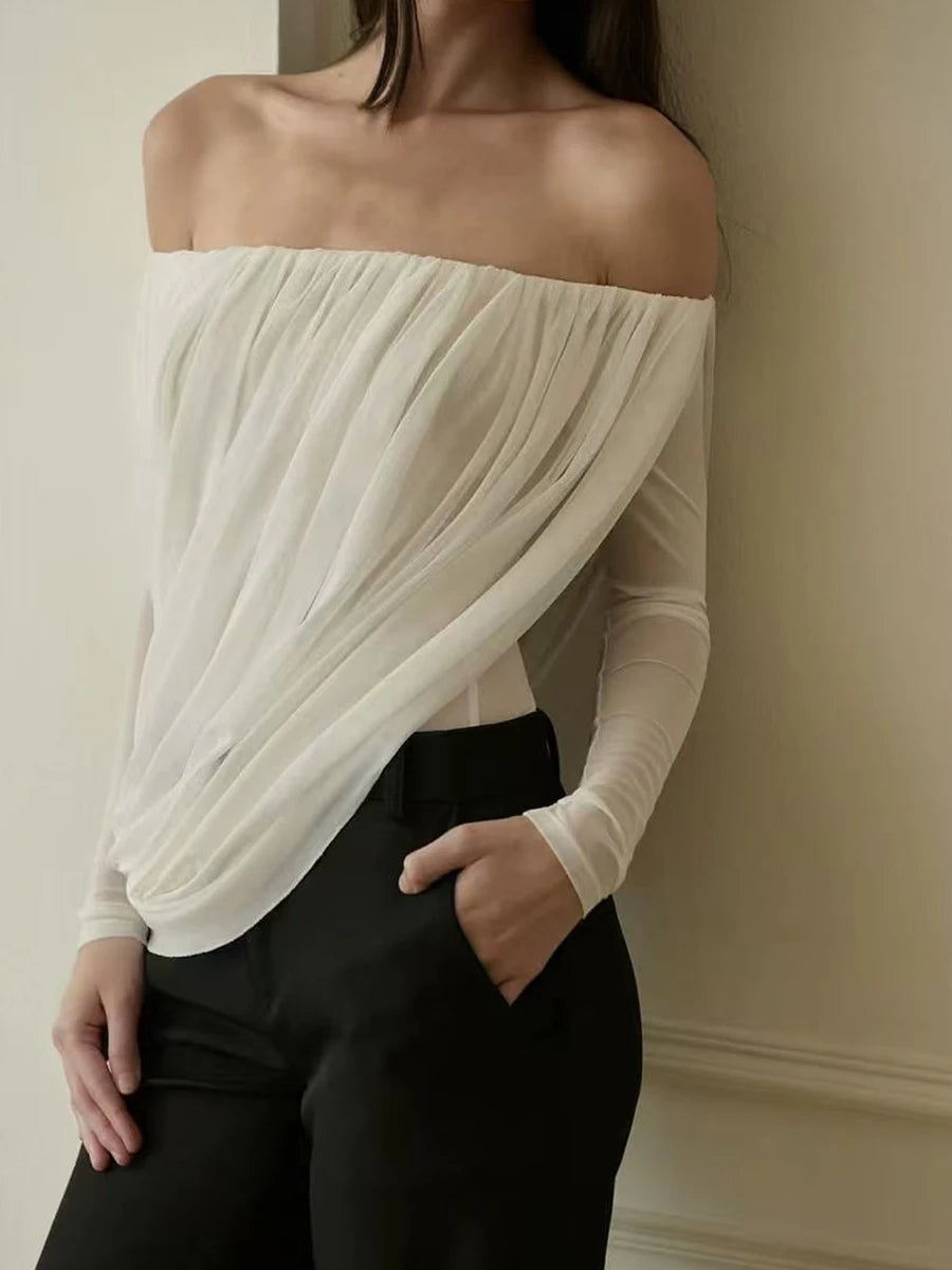 Woman wearing a white off-shoulder top and black pants against a neutral wall.