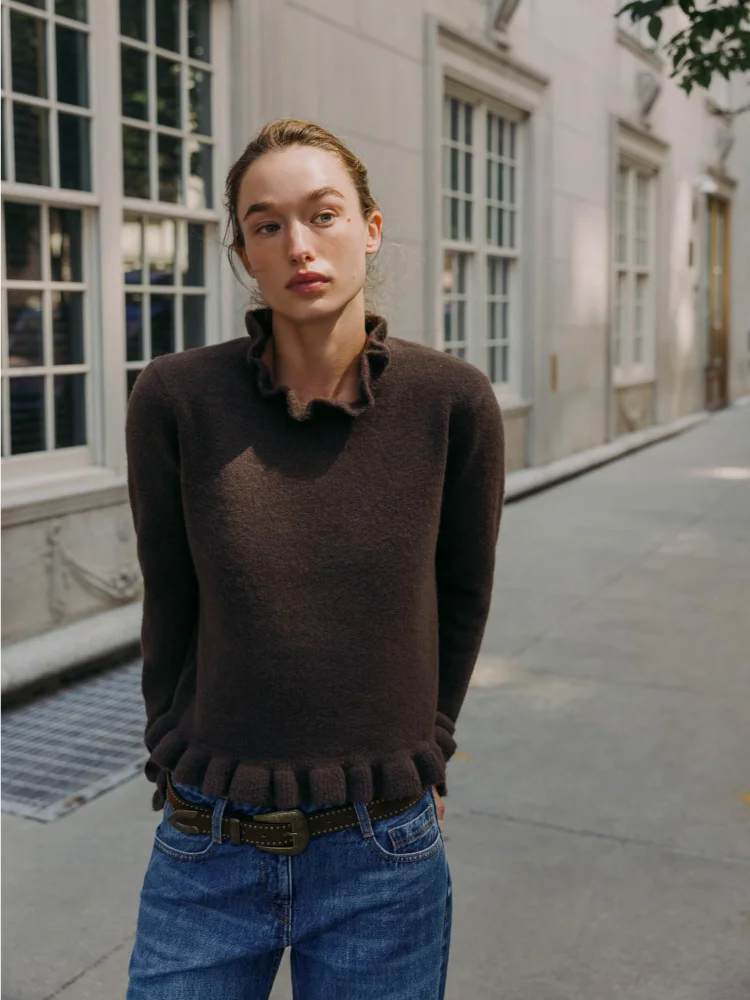 Person wearing a brown sweater and blue jeans standing on a city street.