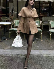 Woman in a beige coat standing in front of a cafe with tables and chairs.