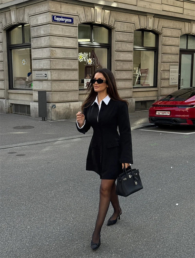 Woman in a black suit walking on a city street with a building and car in the background.