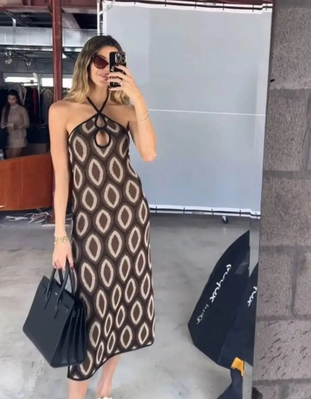 Woman taking a mirror selfie wearing a patterned dress and holding a black handbag.