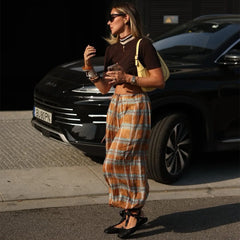 Woman standing next to a black car wearing a brown top and plaid pants.