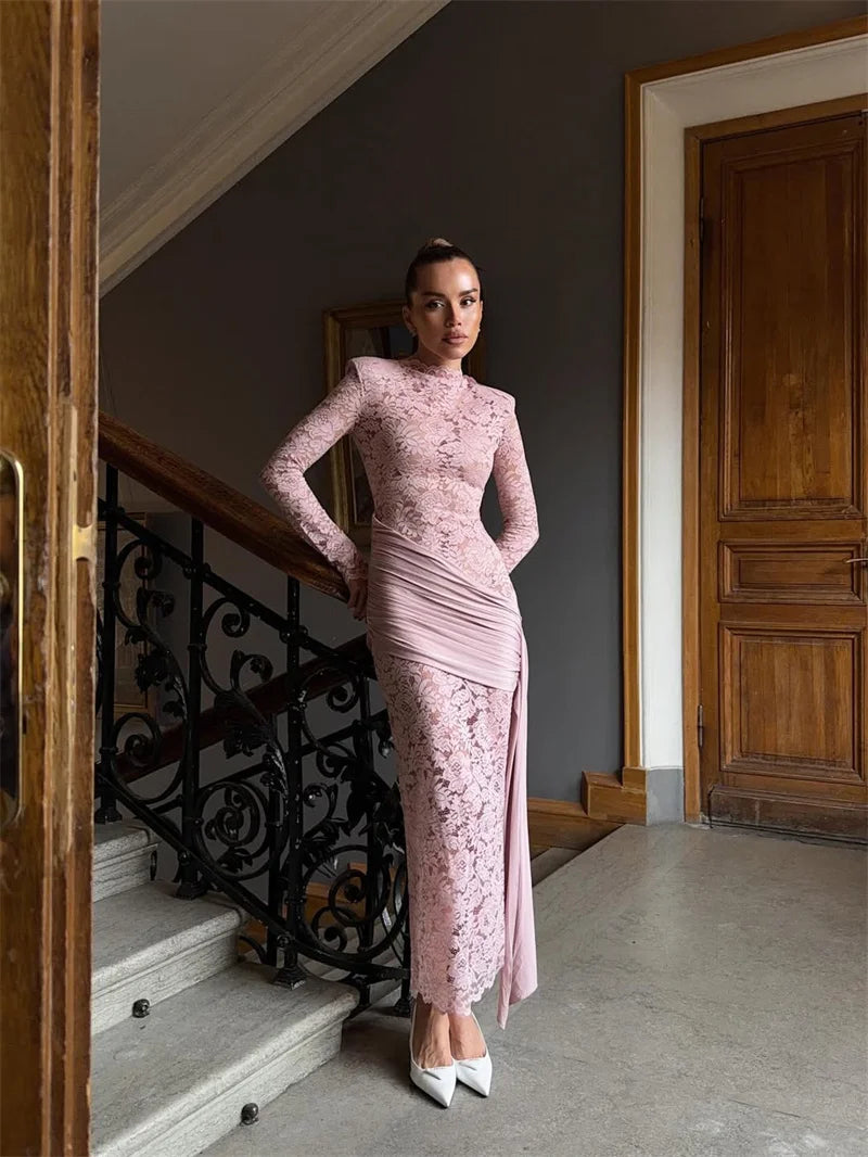 Woman in a pink lace dress standing in an elegant interior setting with wooden doors and a staircase.