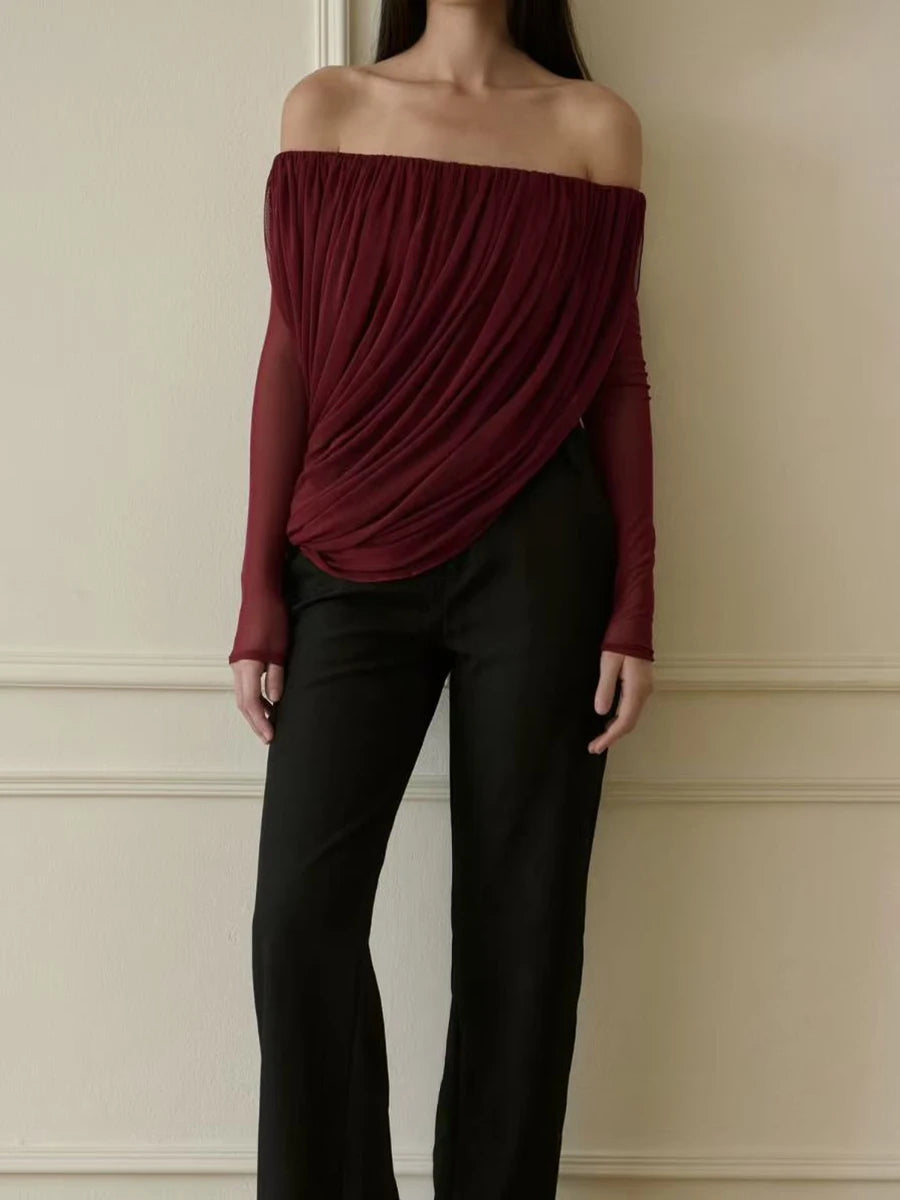 Person wearing a burgundy off-shoulder top and black pants against a beige wall.