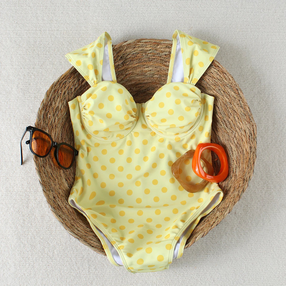 Yellow polka dot one piece swimsuit laid flat in a woven basket with sunglasses and bracelets