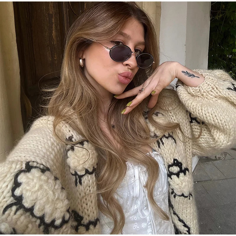 Woman wearing a beige knitted cardigan with black patterns, posing outdoors.