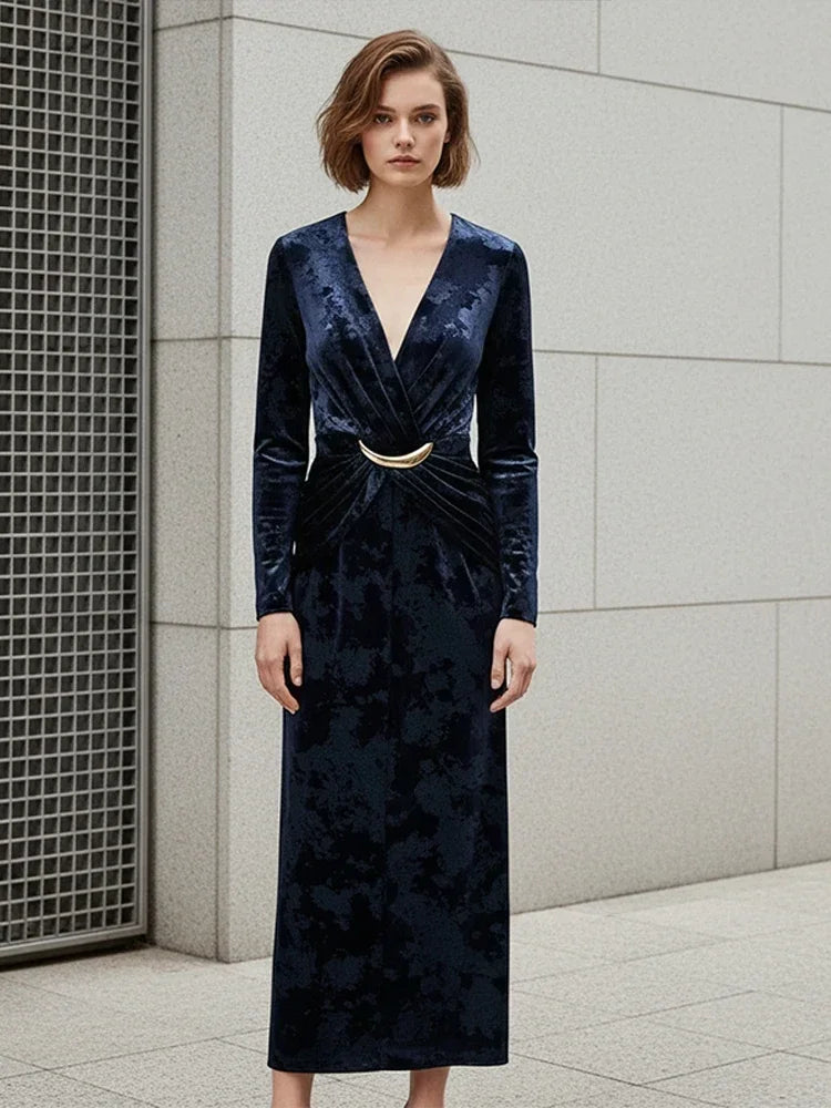 Woman wearing a dark blue velvet dress with a gold belt against a gray tiled wall.