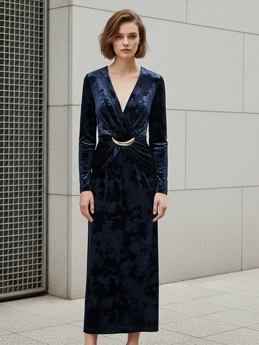 Woman wearing a dark blue velvet dress with a gold belt against a gray tiled wall.
