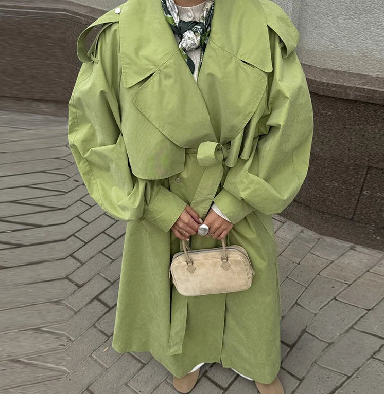 Person wearing a green trench coat holding a beige handbag on a paved surface.
