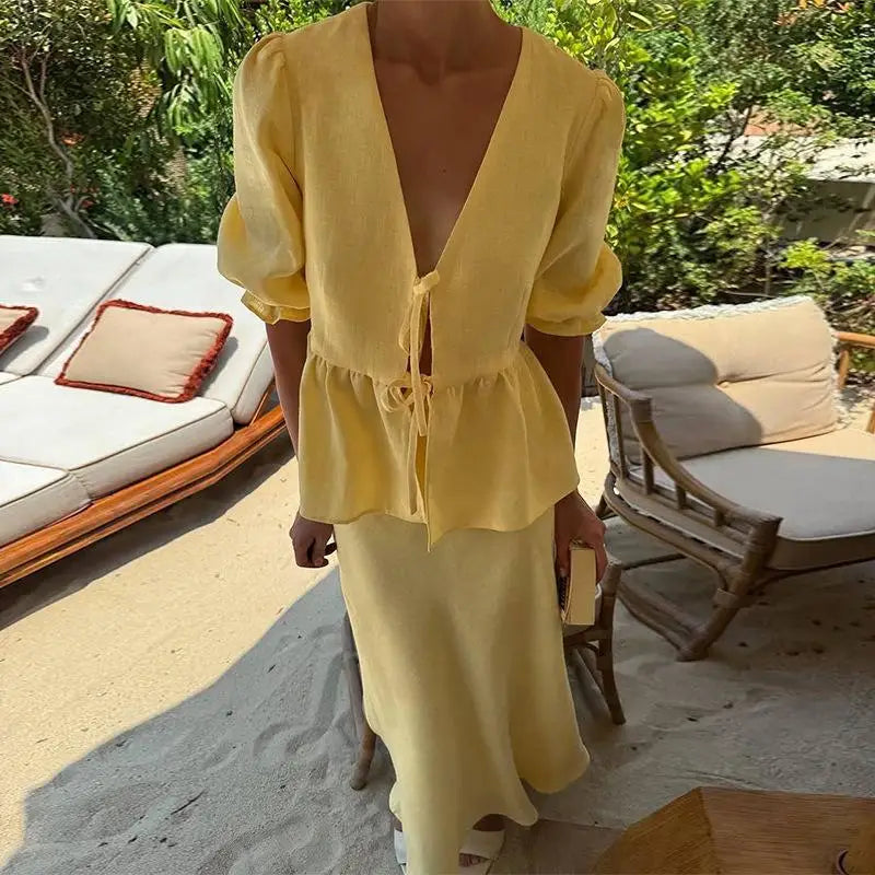 Person wearing a yellow outfit standing on a sandy beach with lounge chairs and greenery in the background.