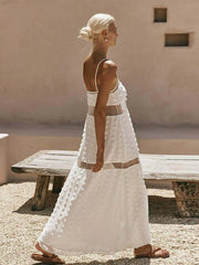 Woman in a white dress standing outdoors with a beige wall and wooden bench in the background.