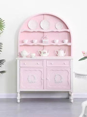 Pink dresser with decorative items in a room with plants and a white chair.