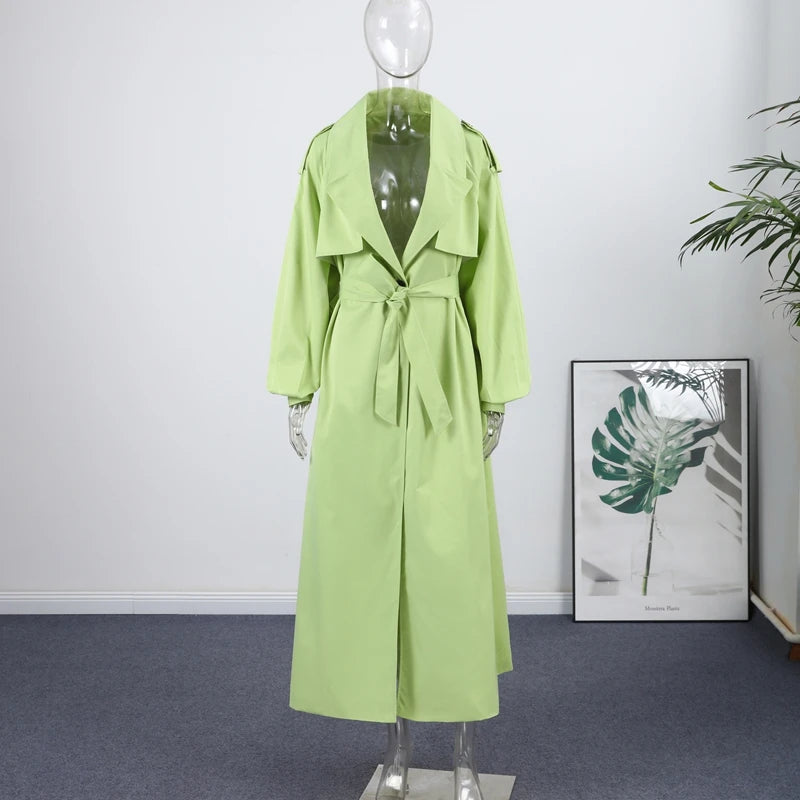 Green trench coat on a mannequin against a white wall with a plant in the corner.