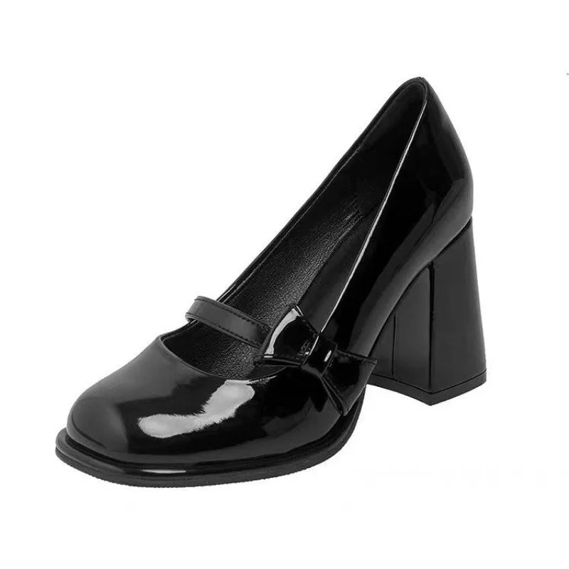 A single black patent leather Mary Jane pump with a chunky block heel and a small bow detail on the strap, shown against a plain white background.
