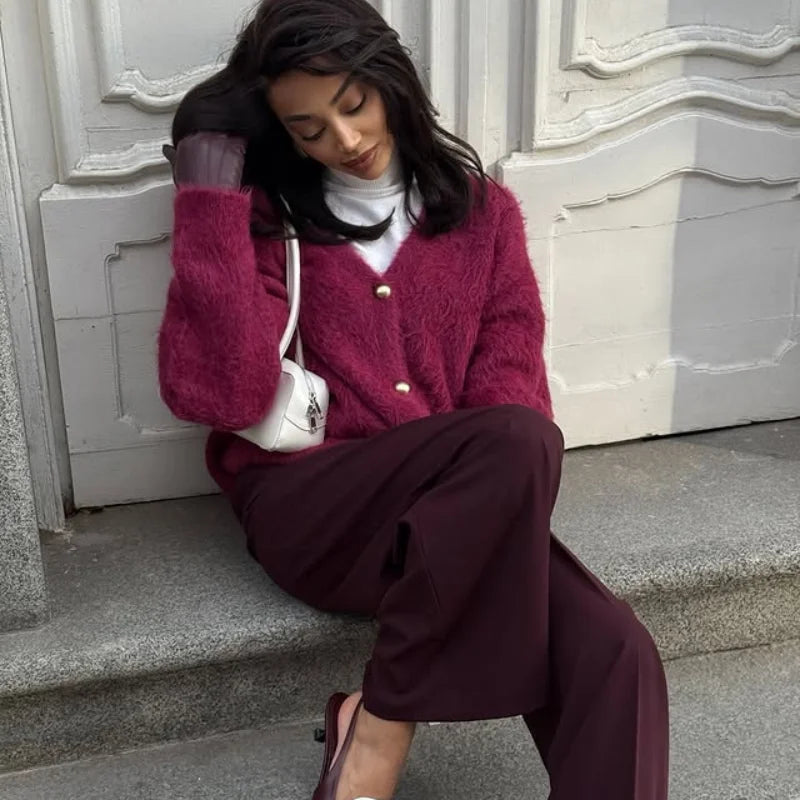 Woman sitting on steps wearing a pink coat and burgundy pants with a white bag.