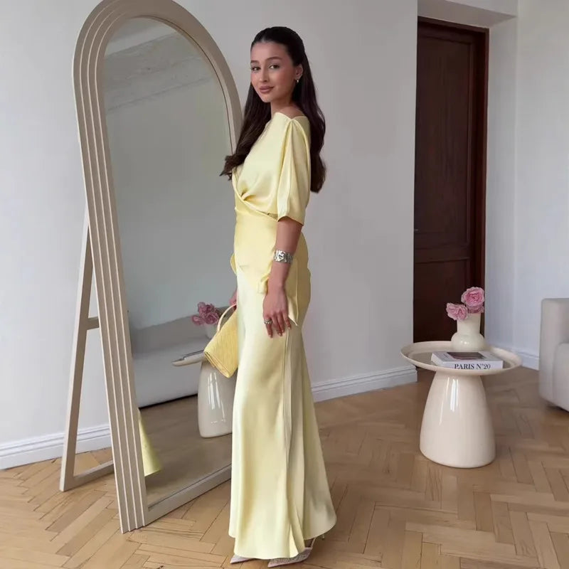 Woman in a yellow dress standing next to a mirror in a room with a table and flowers.