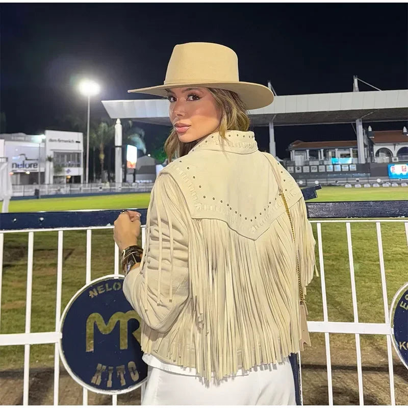 Woman wearing a beige fringe jacket and hat in an outdoor setting with a fence and lights in the background.