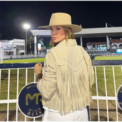 Woman wearing a beige fringe jacket and hat in an outdoor setting with a fence and lights in the background.