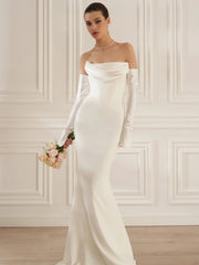 Woman in a white off-shoulder gown holding flowers against a white wall.