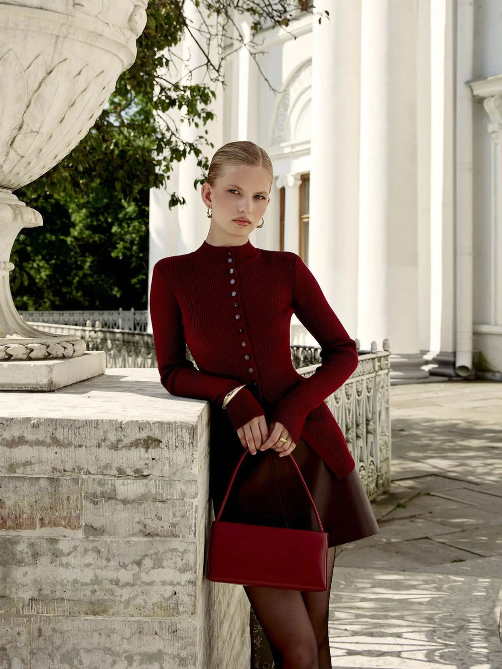 Woman in a red coat and matching handbag standing in an elegant outdoor setting.