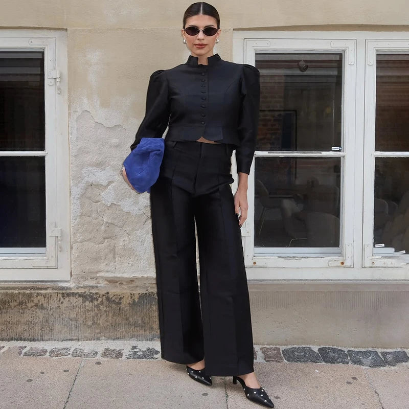 Woman in a black outfit holding a blue clutch against a beige wall with windows.