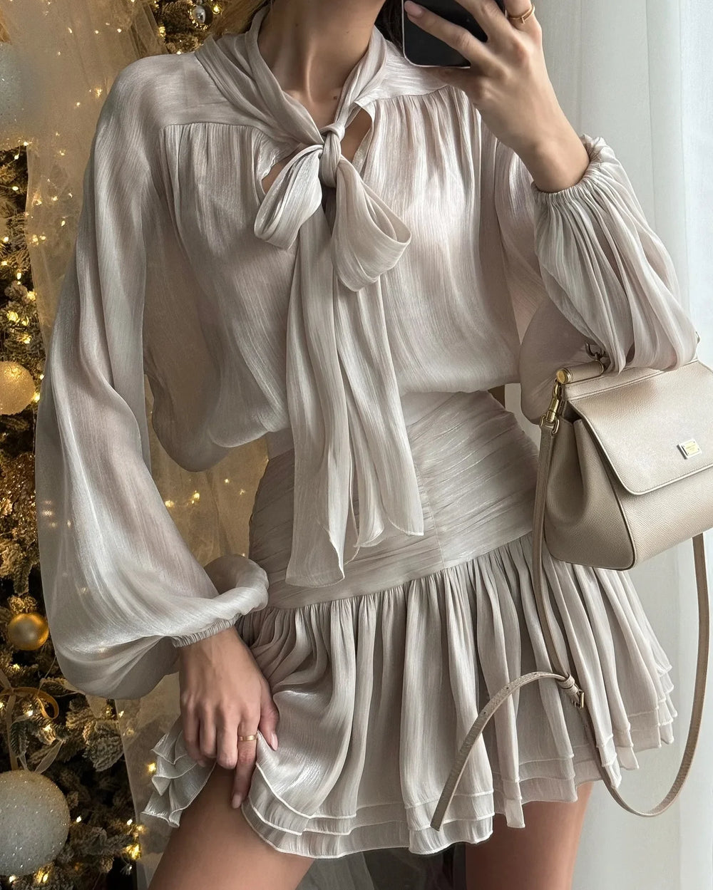 Person wearing a white blouse with a bow and a matching skirt, holding a beige handbag in front of a decorated Christmas tree.