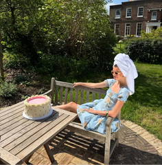 Woman sitting on a bench in a garden with a cake, wearing a blue dress and towel on her head.