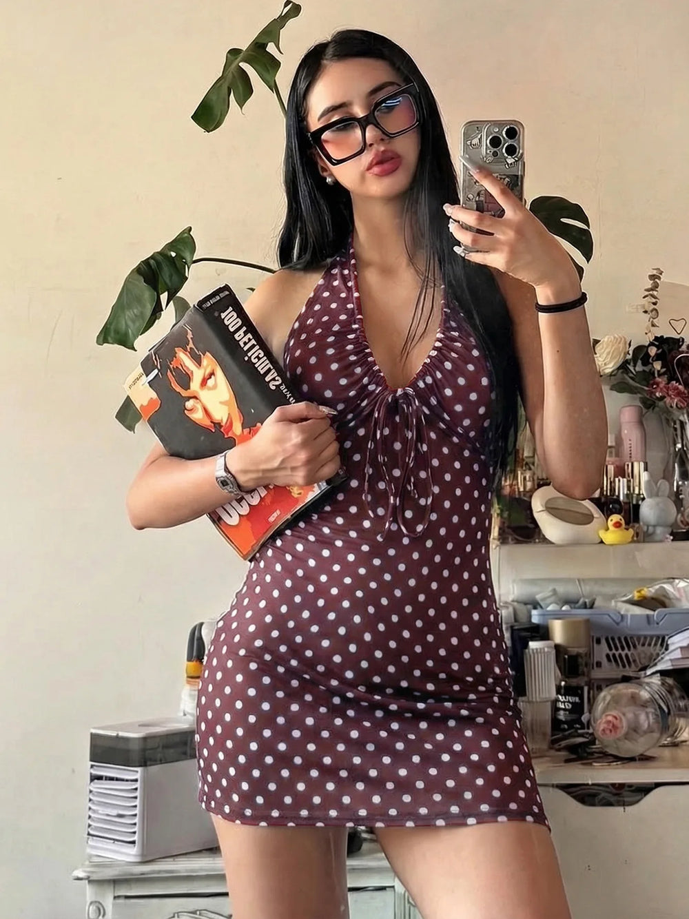Woman in a polka dot dress holding a book in a room with decor elements.