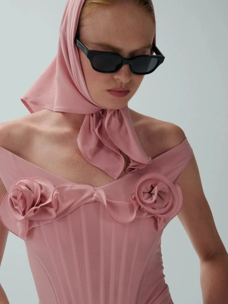 Woman wearing a pink dress with floral details and a matching headscarf against a plain background