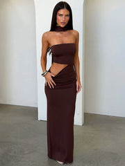 Woman wearing a brown strapless maxi dress with a side cutout and halter neck detail, posing against a white wall.