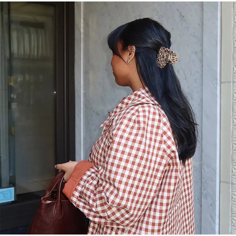 Woman in a red and white checkered shirt with a brown handbag, standing against a gray wall.