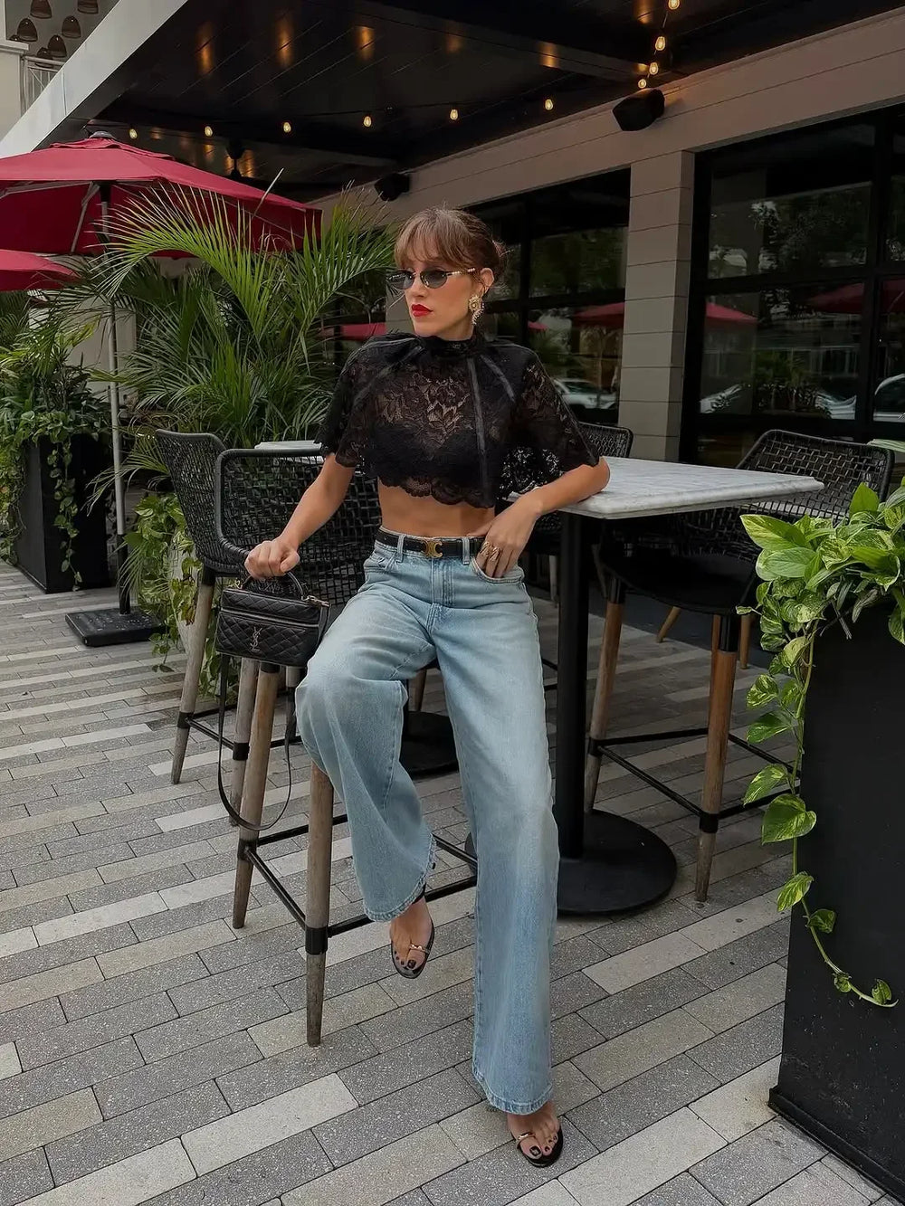 Woman sitting on a stool in an outdoor setting with plants and tables.