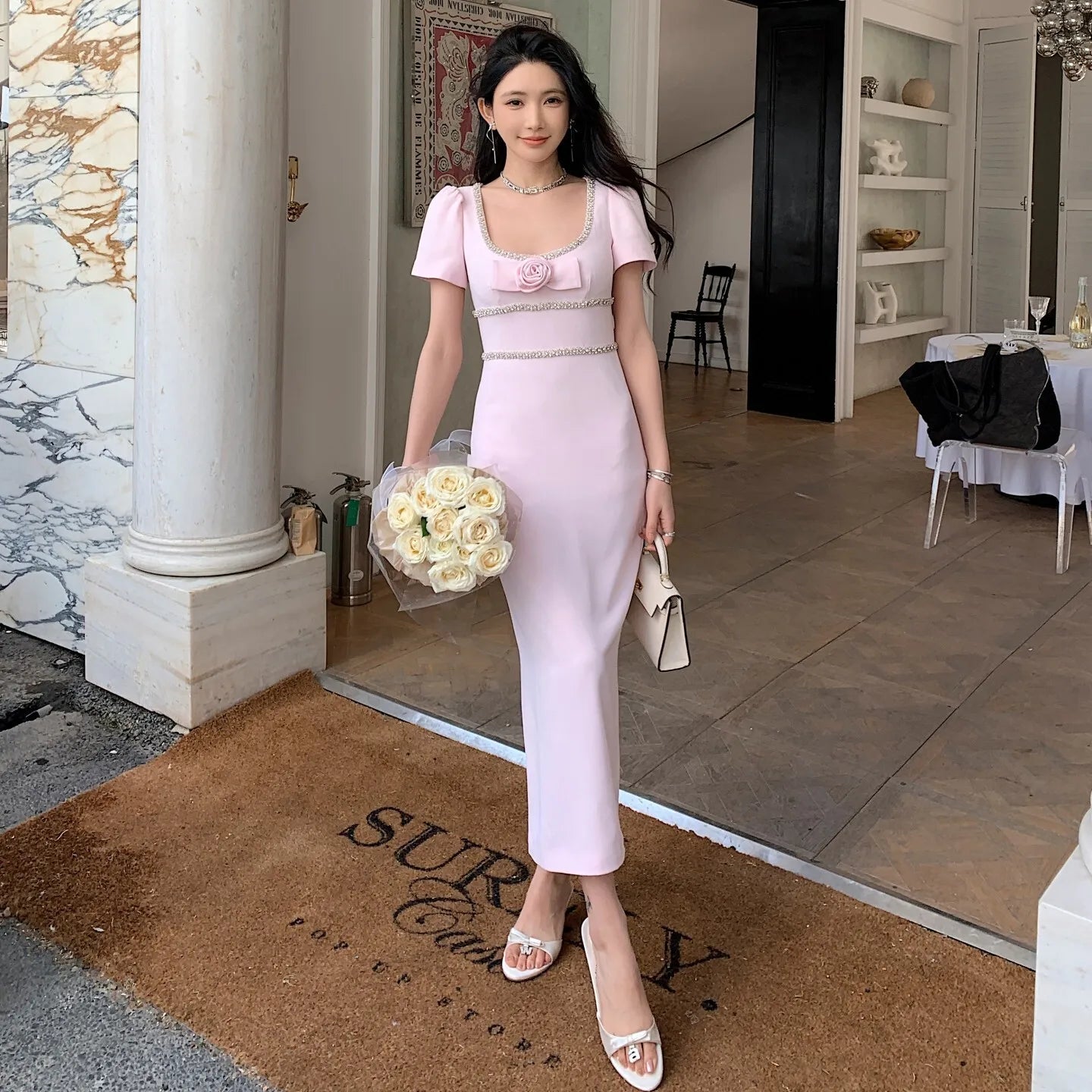 Woman in a pink dress holding flowers on a doormat with 'Surya' branding.