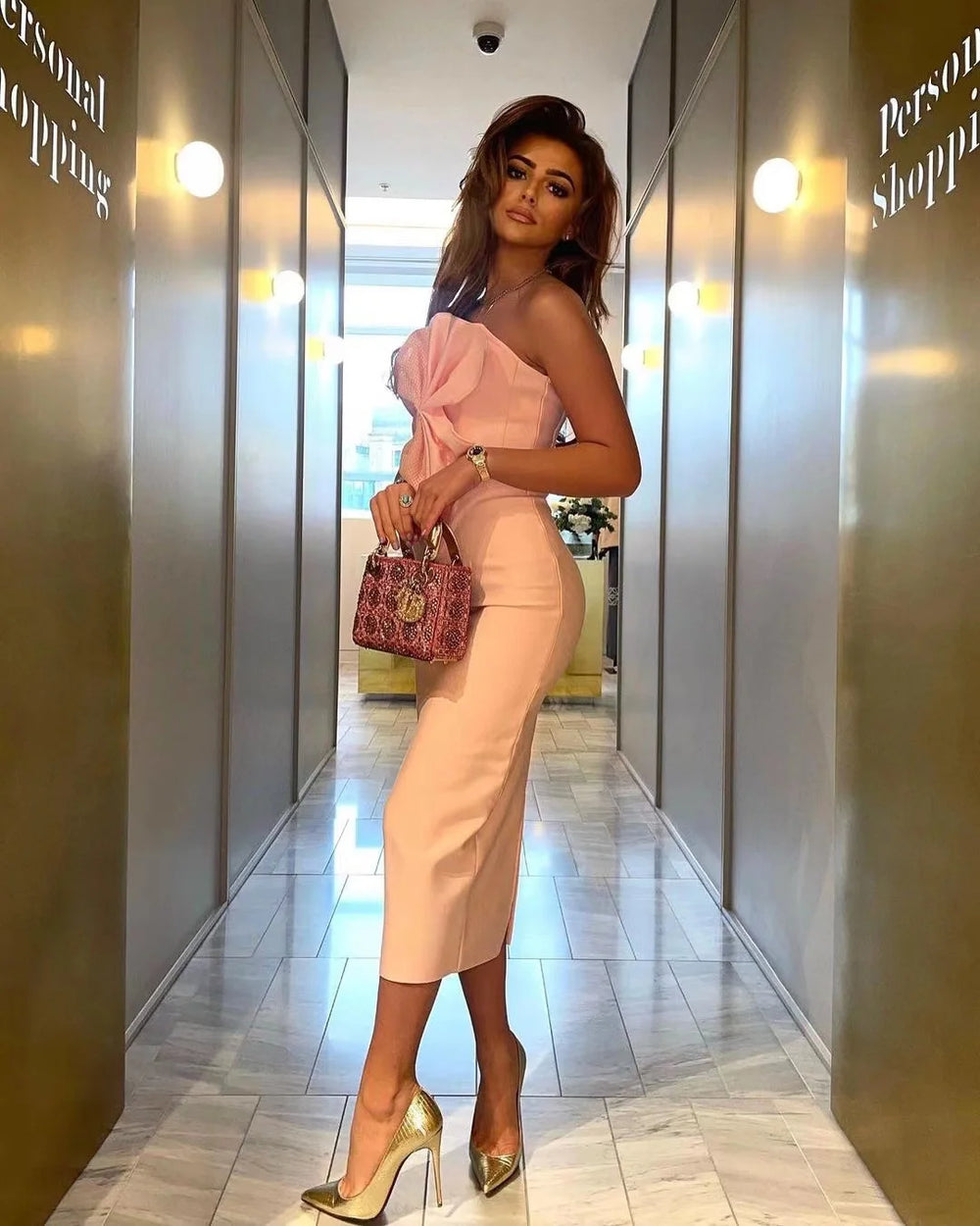 Woman in a pink strapless dress and gold high heels standing in a hallway.