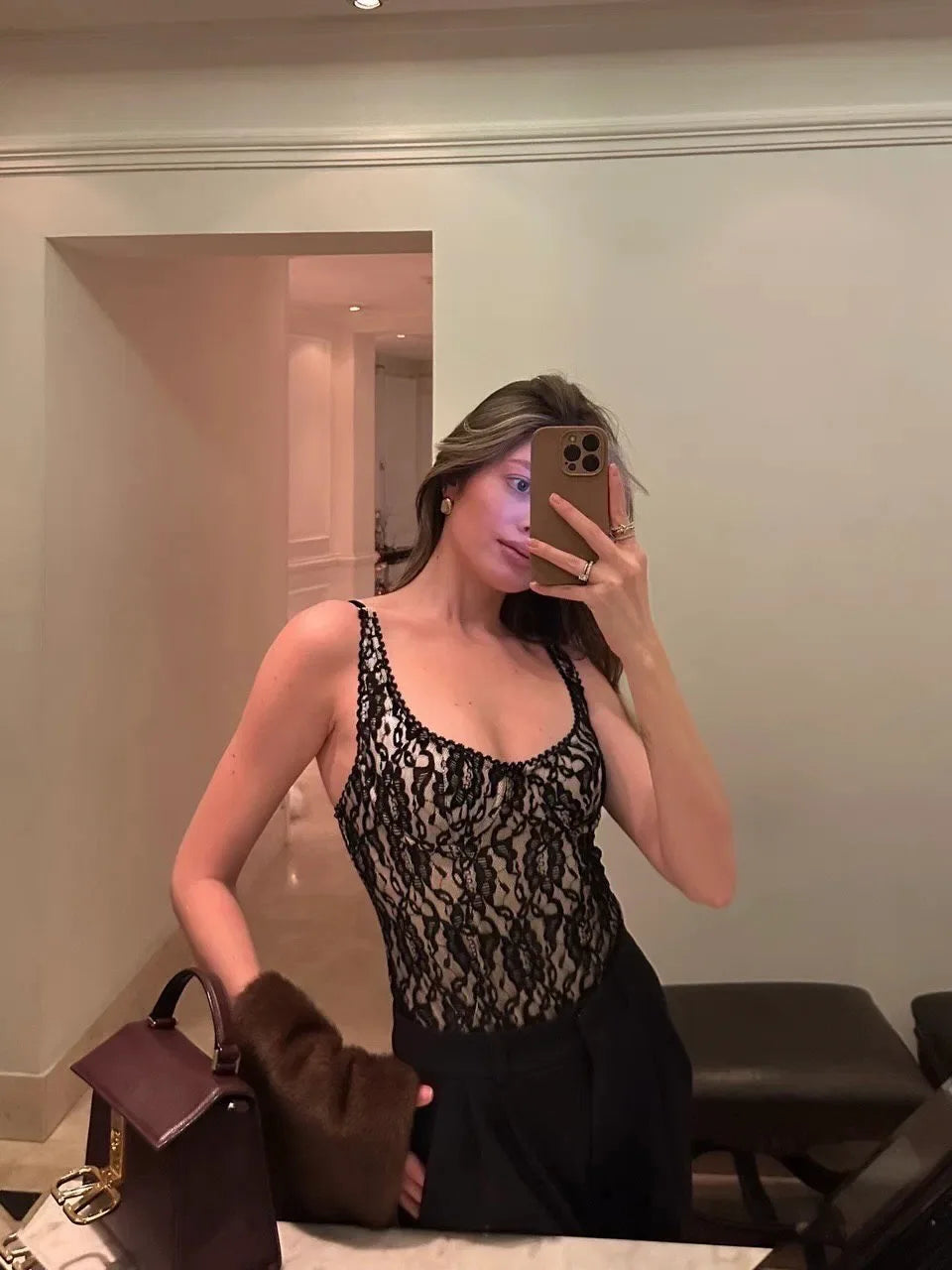 Woman taking a mirror selfie wearing a black lace top and holding a brown handbag.
