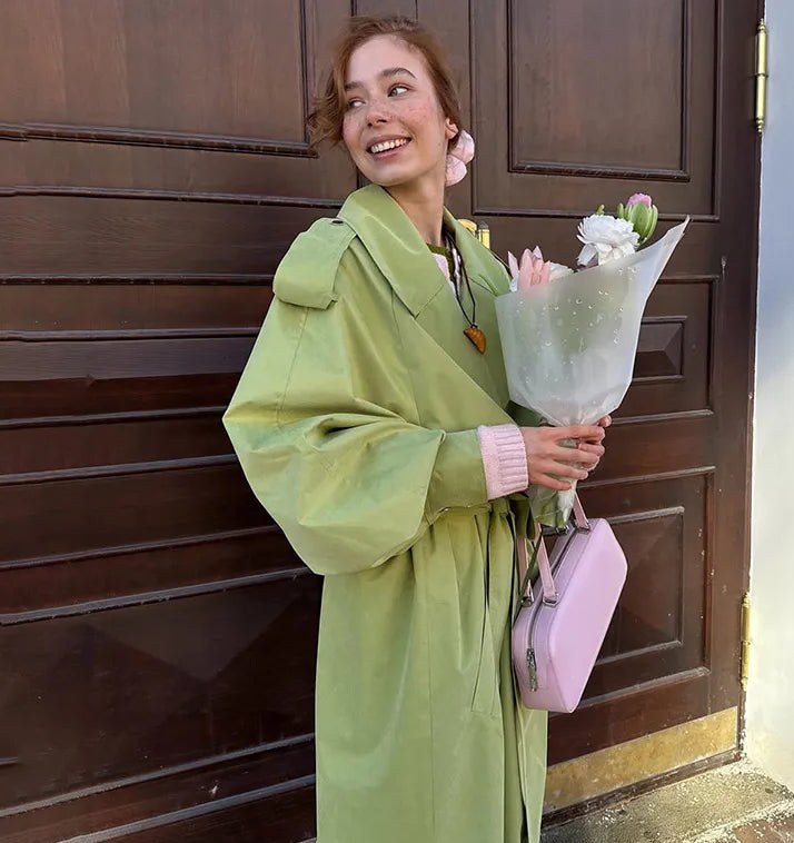 Woman in a green coat holding flowers and a pink bag against a wooden door.