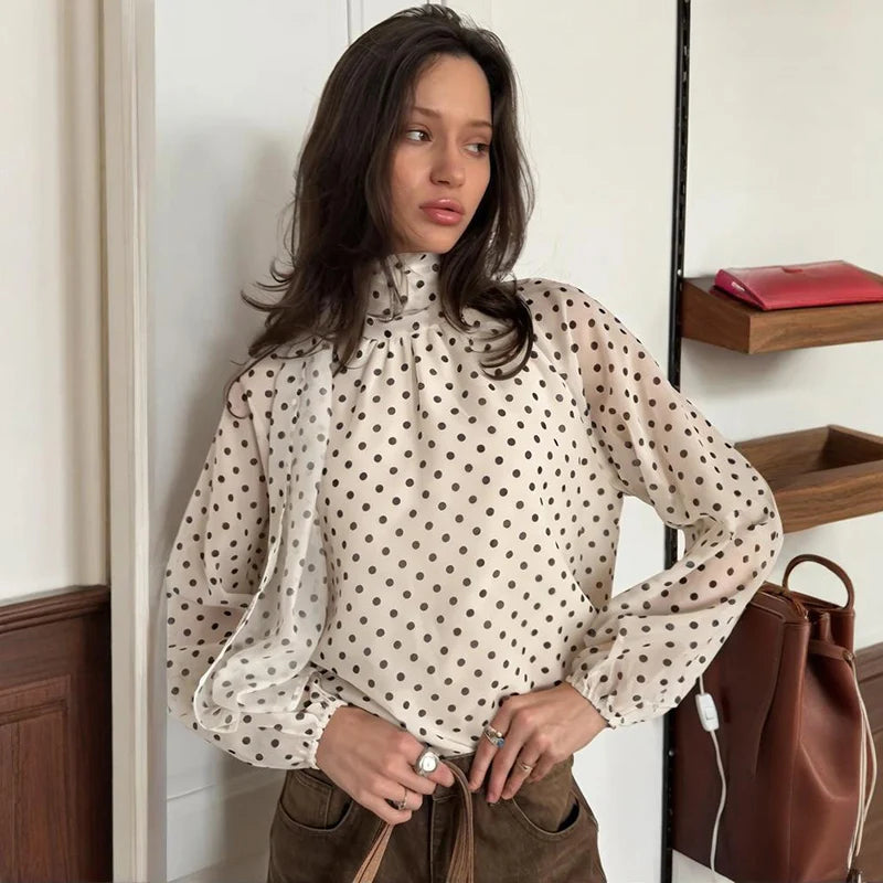 Woman wearing a polka dot blouse standing indoors with a handbag nearby.