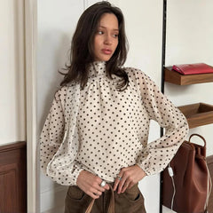 Woman wearing a polka dot blouse standing indoors with a handbag nearby.