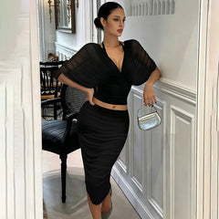 Woman leaning against wall in black V-neck draped sleeve crop top and ruched midi skirt set