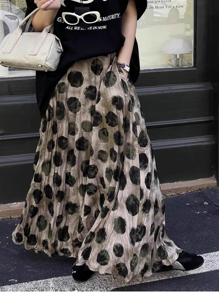 Person wearing a black t-shirt with text and a long polka dot skirt, standing on a street.